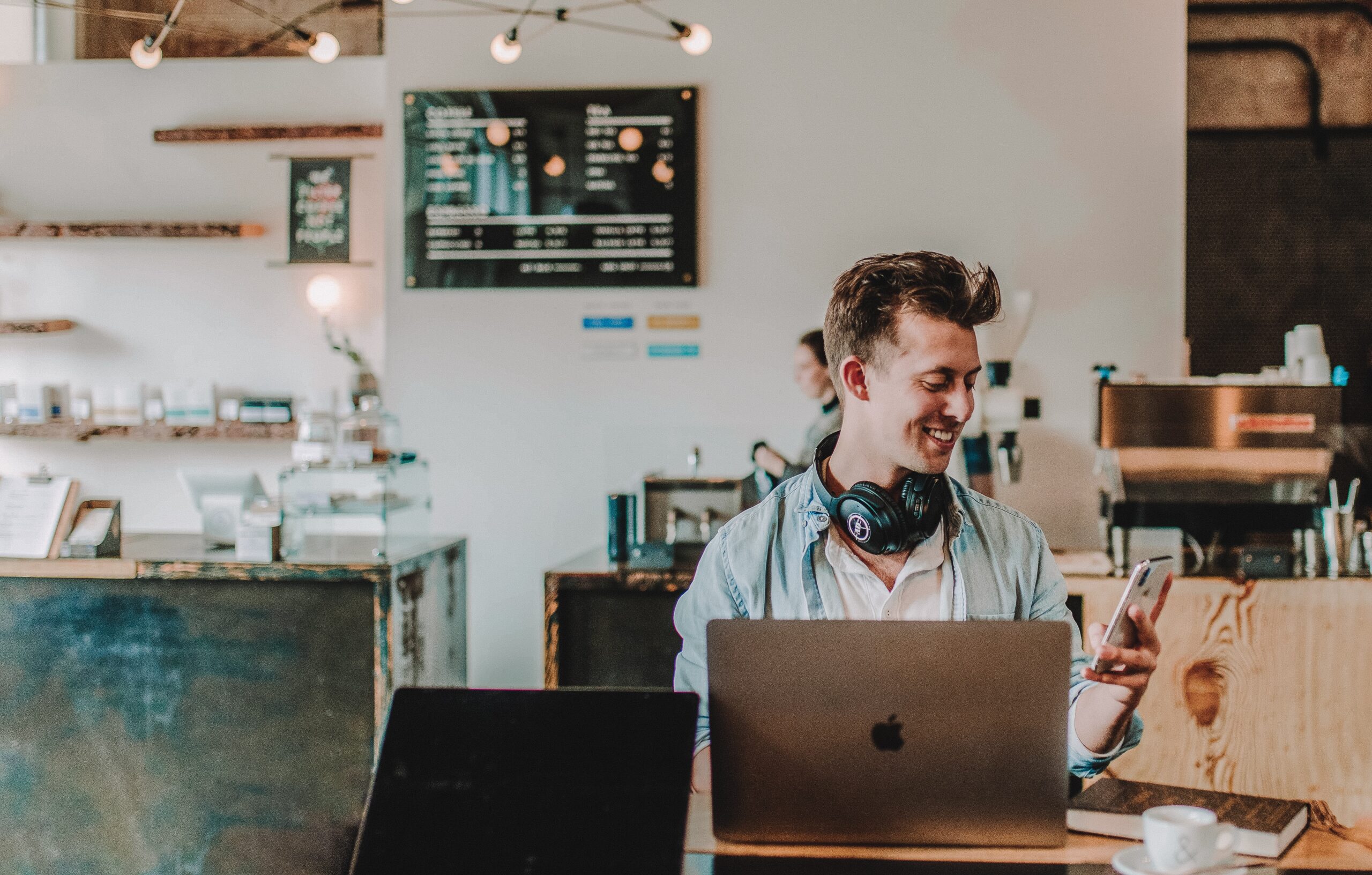 app developers A man with headphones around his neck is smiling while looking at his phone, likely using an app, seated at a table with a laptop in a café environment. tiny screen labs