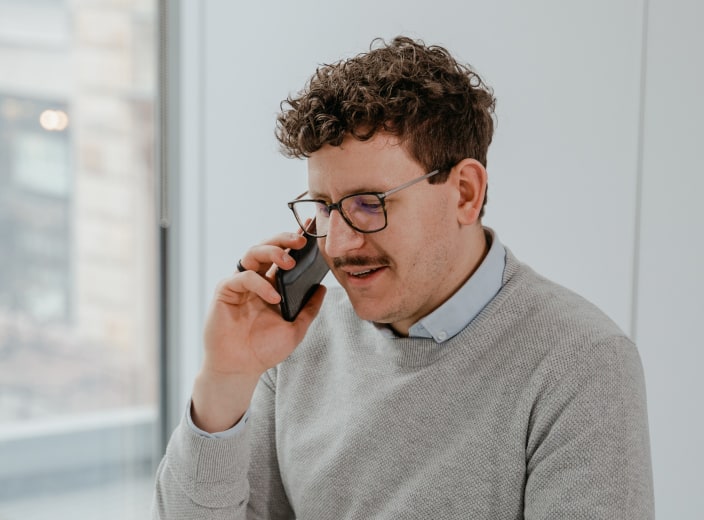 app developers man with glasses and curly hair, wearing a gray sweater, talking on a smartphone in a bright room. tiny screen labs